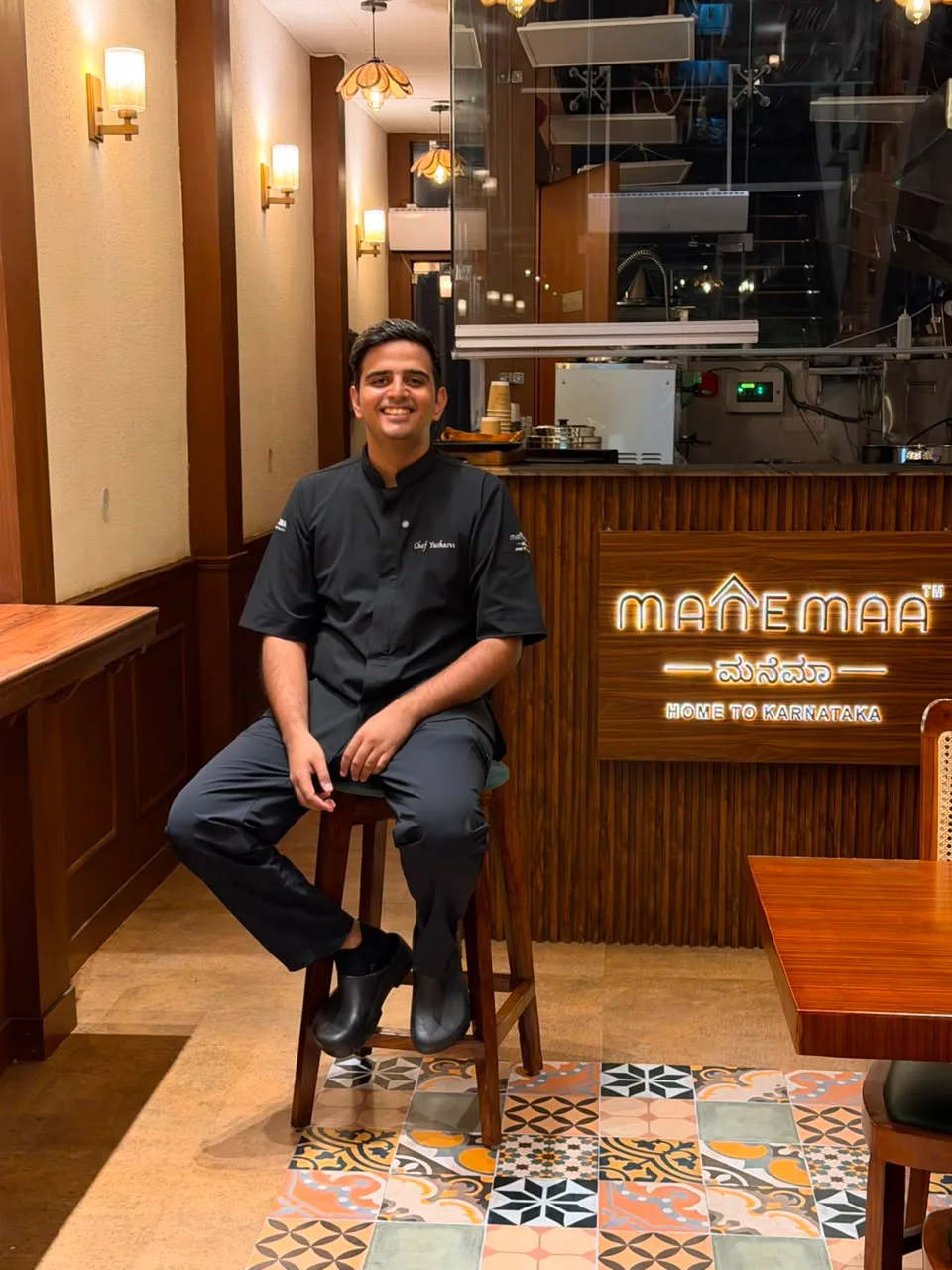 Yashasvi Yadav, chef and founder, in his chef’s jacket inside the MANEMAA dining room, with the illuminated MANEMAA sign behind him